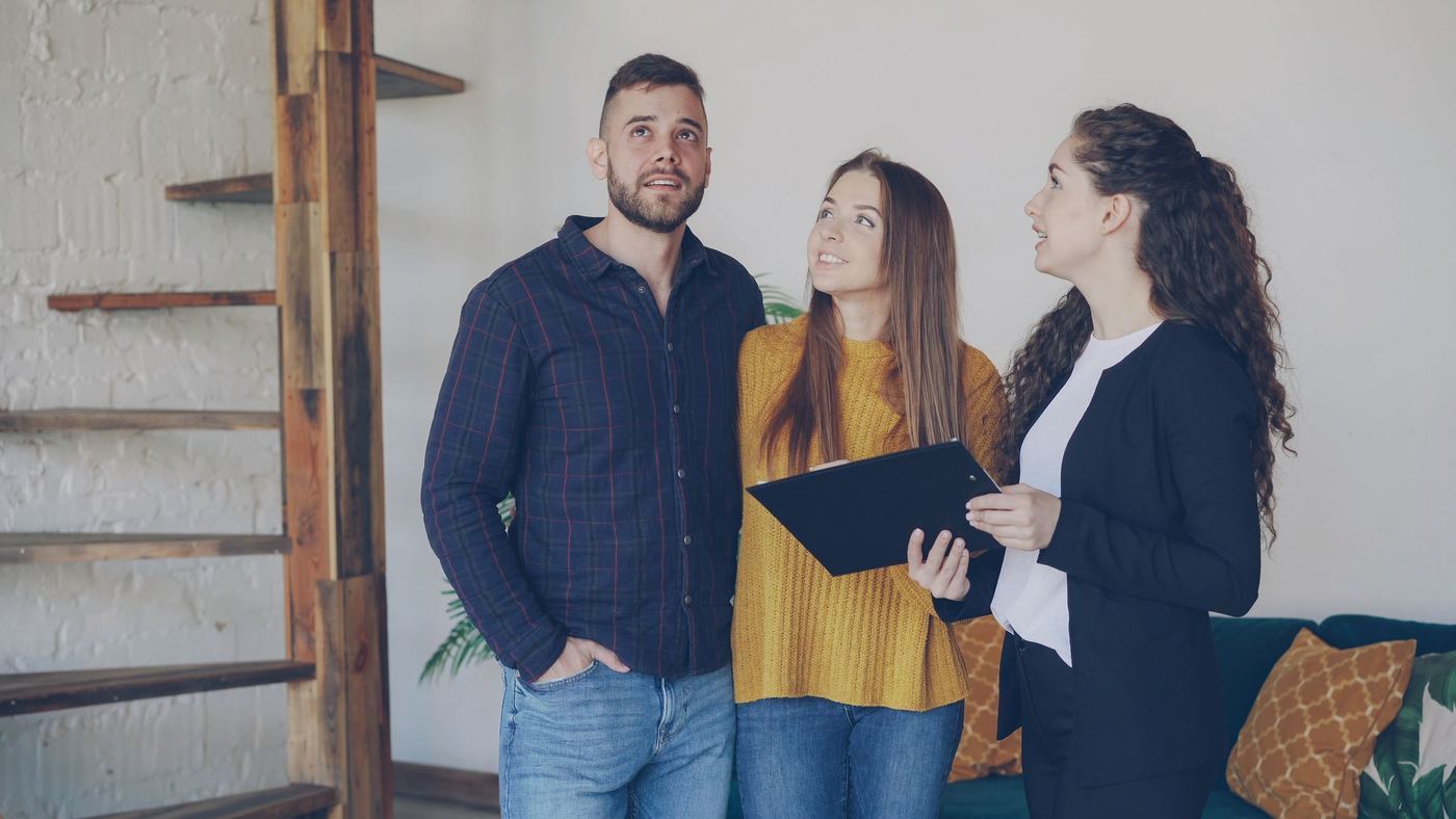 A young couple touring a home with a real estate agent reviewing a clipboard indoors